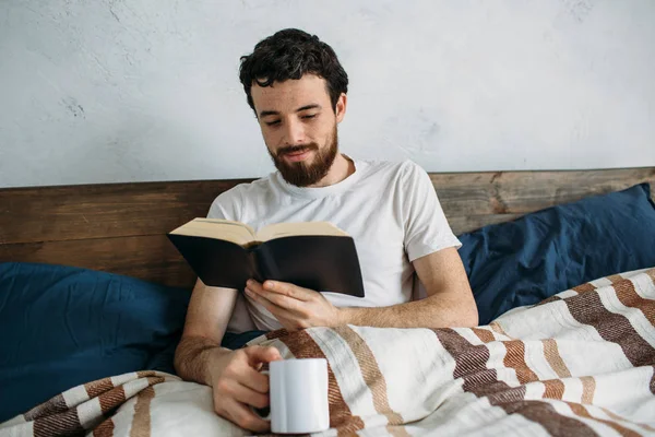 Bärtiger Mann liest ein großes Buch, das in seinem Schlafzimmer liegt. — Stockfoto