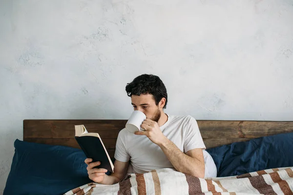 Bärtiger Mann liest ein großes Buch, das in seinem Schlafzimmer liegt. — Stockfoto