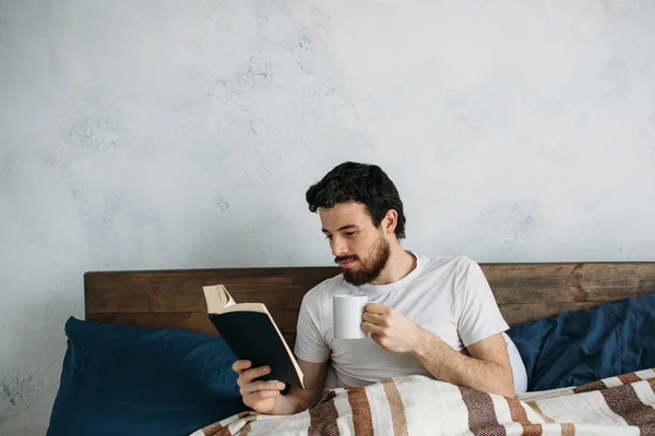Bärtiger Mann liest ein großes Buch, das in seinem Schlafzimmer liegt. — Stockfoto