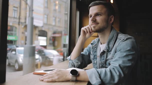 Счастливчик сидит в кафе рядом с окном с блокнотом — стоковое видео