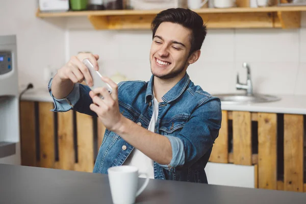 Joven sentado en una mesa en la cocina con su teléfono móvil —  Fotos de Stock