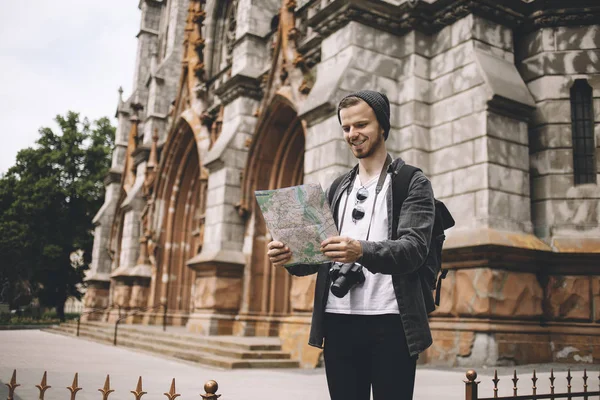 Young and ambitious tourist is standing on the sidewalk near the cathedral and reading a map