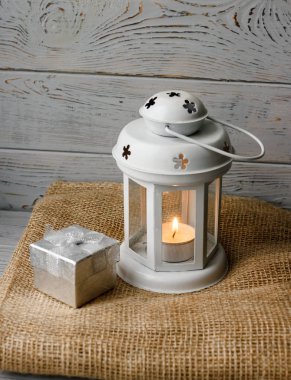 Kahverengi masa örtüsü içinde yanan bir mum ile beyaz fener. Dekoratif yıldız şamdan. Ahşap arka plan. Mumlar yanında gümüş hediye kutusu.