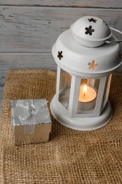 Kahverengi masa örtüsü içinde yanan bir mum ile beyaz fener. Dekoratif yıldız şamdan. Ahşap arka plan. Mumlar yanında gümüş hediye kutusu.