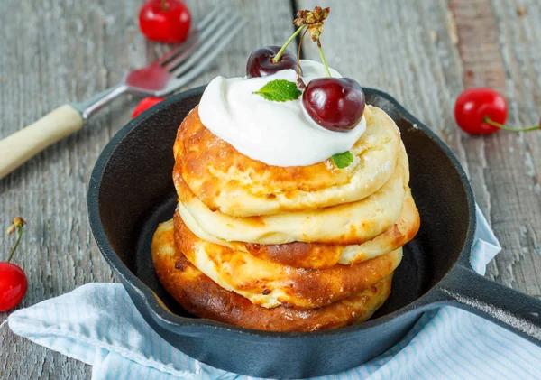 Homemade cottage cheese pancakes with sour cream and cherries in a cast-iron frying pan.