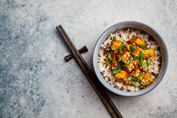 Marinated tofu with sesame seeds, green onions and brown rice in a bowl on a gray background. Asian food concept.