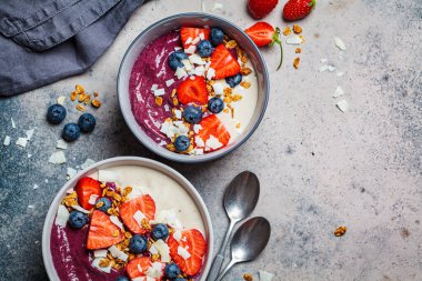 İki renkli meyve suyu kasesi, granola, yabanmersini ve hindistan cevizi, gri arka plan. Sağlıklı vejetaryen gıda kavramı.