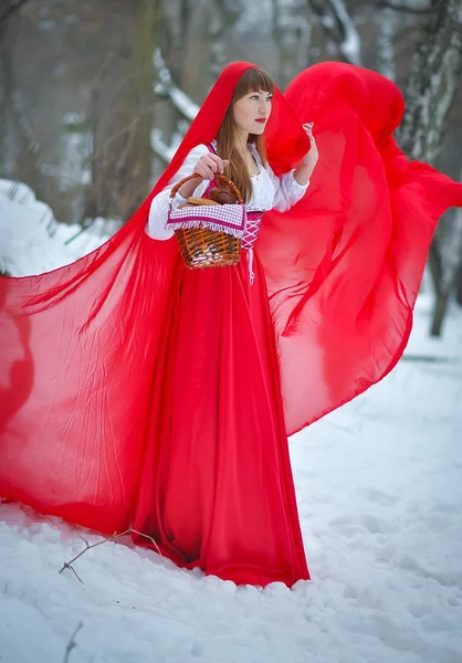 Mujer capucha roja de pelo rubio con una larga capa roja y vestido rojo viejo camina en el bosque de invierno, Una encantadora joven, entre árboles y nieve, sosteniendo un pastel con cesta, cuentos de hadas — Foto de Stock