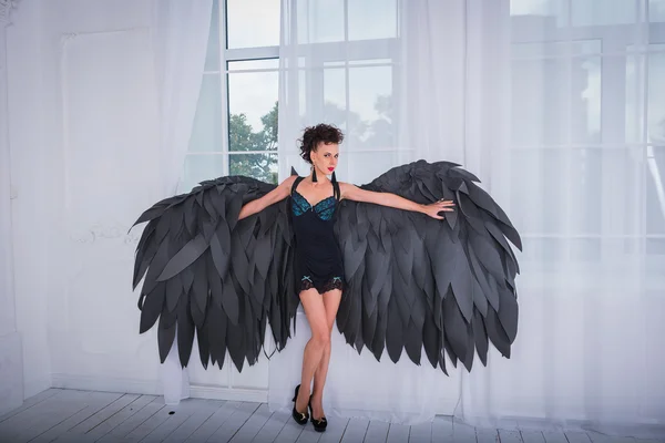 Femme brune ange noir avec les grandes ailes, lèvres rouges, costume court noir, dans la chambre blanche debout près de la fenêtre Image En Vente