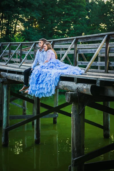 Una coppia di storie d'amore, innamorati, insieme nel parco forestale, seduti sulla fine del ponte di legno, ragazza in un bellissimo vestito viola, serata soleggiata, estate, acqua verde sullo sfondo Immagini Stock Royalty Free