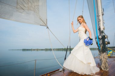uzun beyaz bir elbise içinde güzel sarışın gelin bir yelkenli yat ile mavi buket denizde üzerinde elini ve ipi Öte yandan güneşli bir günde poz veriyor 