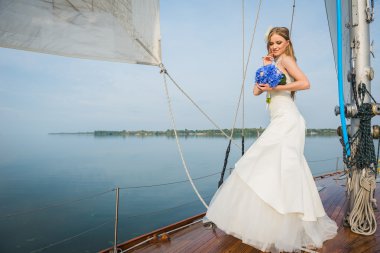 uzun beyaz bir elbise içinde güzel sarışın gelin bir yelkenli yat ile mavi buket denizde üzerinde onun elinde güneşli bir günde poz veriyor 