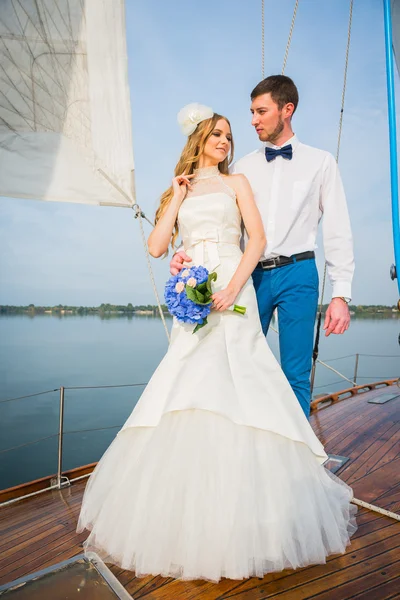 Boldog menyasszony és a vőlegény, átölelve egy vitorlás jachton a kék csokrot a kezében, egy napsütéses napon; elegáns, hosszú ruha, csokornyakkendő — Stock Fotó