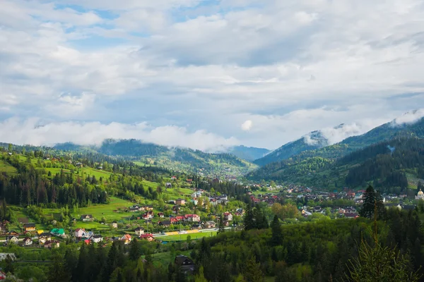 Primavera nelle montagne dei Carpazi, Ucraina, mattina di sole, vista dall'alto, una piccola città e colline forestali sul paesaggio, cielo blu e nuvole bianche, un po 'di nebbia sulla cima della collina Immagine Stock