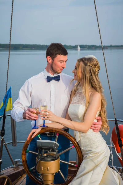 Boldog menyasszony és a vőlegény, átölelve egy vitorlás jachton pezsgővel a kezében, egy napsütéses napon; elegáns, hosszú ruha, csokornyakkendő — Stock Fotó