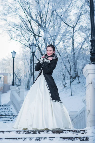 Portrait d'hiver d'une mariée en robe blanche debout sur les escaliers - la neige est partout, les arbres sous la neige à l'arrière-plan et les lumières vintage Photos De Stock Libres De Droits
