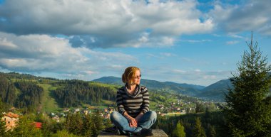 bir kadın oturmuş, yüksek dağlarda dinleniyor. güneşli bir gün, Bahar, mavi gökyüzü ve arka plan ve yüksek küçük bir kasabadan beyaz bulutlar üzerinde yukarıda. bir güneş gözlüğü ve kot pantolon