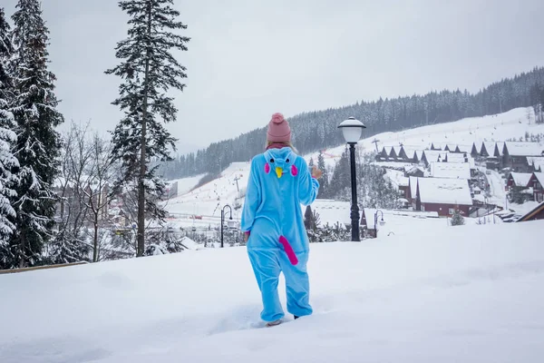 Dziewczyna w niebieski, Różowy Jednorożec pijama kigurumi odkryty przed drewna domy w raporcie dla narciarzy w górach śniegu. — Zdjęcie stockowe