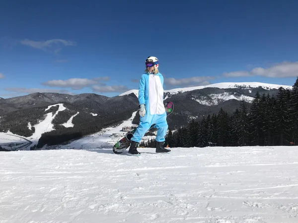 Girl in blue, pink unicorn pajama kigurumi outdoor with snowboard on the ski report in snow mountains. — Stock Photo, Image