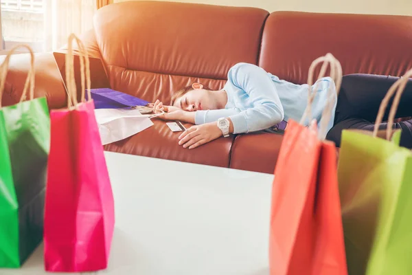 Mujer joven infeliz dormir en el sofá y mantener bolsas de compras con de —  Fotos de Stock