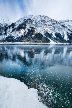 Karla kaplı dağlar göle yansıyor, çok kar yağıyor. Güzel kış manzarası, parlak renkler. 