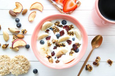 Meyve ve meyveli bir kase sağlıklı yoğurt, taze muz, kurutulmuş meyveler, beyaz ahşap arka planda bir fincan sade kahveyle servis edilir.. 
