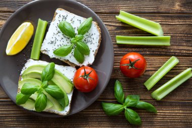 Sağlıklı avokado sandviçleri, krem peynir, taze kereviz çubukları, kiraz domatesleri ve fesleğen yaprakları, üst manzara, kahverengi masa arka planı. Bir dilim sarı limon. Yakın plan.. 
