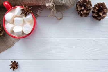 Şekerlemeli sıcak kakao, kırmızı bardak ve çuval bezi, kozalak, anason yıldızı süslemesi. Metin için boş alan. Beyaz tahta masa arkaplanı. 