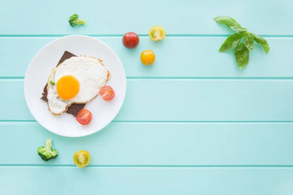 Rye bread toasts with fried eggs and vegetables: cherry tomatoes, broccoli and fresh basil leaves on turquoise table background. Copy space, space for text.