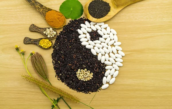 Beras hitam dan pil putih membentuk simbol yin yang dan bola kompresi herbal Spa, bubuk kunyit, millet, kedelai, biji kemangi dalam sendok kayu pada campuran kayu cokelat herbal dan obat-obatan Stok Foto Bebas Royalti