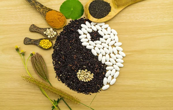 Black rice and white pill forming a yin yang symbol and Spa herbal compressing ball , turmeric powder , millet , soybean , basil seed in wooden spoon on brown wooden blending of herb and medicine — Stock Image Black rice and white pill forming a yin yang symbol and Spa herbal compressing ball , turmeric powder , millet , soybean , basil seed in wooden spoon on brown wooden blending of herb and medicine Stock Photo