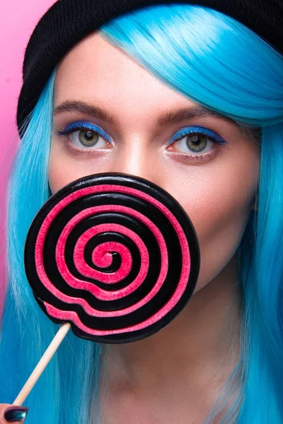 Beauty portrait of a young woman with lollipop with cyan hair and creative makeup on pink background in studio