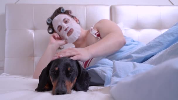 Metrosexual guy with hair curlers and fabric mask for smoothing wrinkles on face lies on bed at home or in hotel and strokes sleeping dachshund dog. Handsome man caring for himself before event. — Stock Video