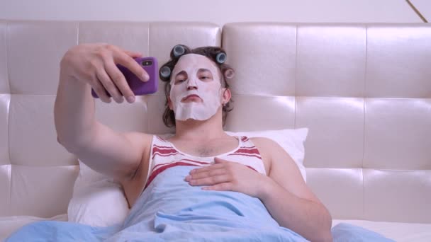 Grappige man blogger met een cosmetisch masker en krulspelden in een kamer maakt selfie foto — Stockvideo