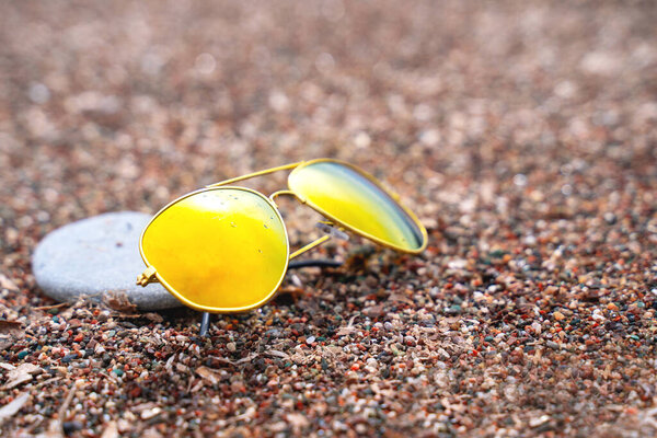 Sunglasses with polarizing lenses with pebble nearby lie on sand, close up. Protection against harmful ultraviolet radiation from sun. Accessories for vacation trip to warm countries to sea or ocean.