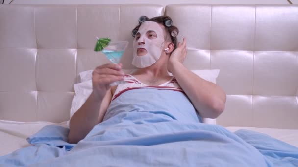 Man with a cosmetic mask makes spa treatments, holding a cocktail make any faces — Stock Video