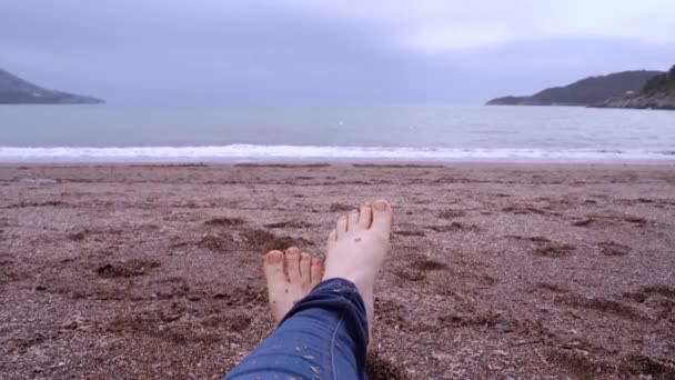 Ludzkie nogi w dżinsach leżą na mokrym piasku na plaży, palce się poruszają. Niespokojny krajobraz morski z górami w pobliżu horyzontu w pochmurny dzień. Podróżowanie samemu, czas wolny na samotność, medytację i planowanie na przyszłość. — Wideo stockowe