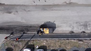 Asfalt sıkıştırma silindiri yeni kaldırımı küçültür ve düzleştirir, yükseklikten üst görünüm. Kürekli inşaat işçileri süreci izliyorlar. Yolların ve otoyolların yapımı ve onarımı.