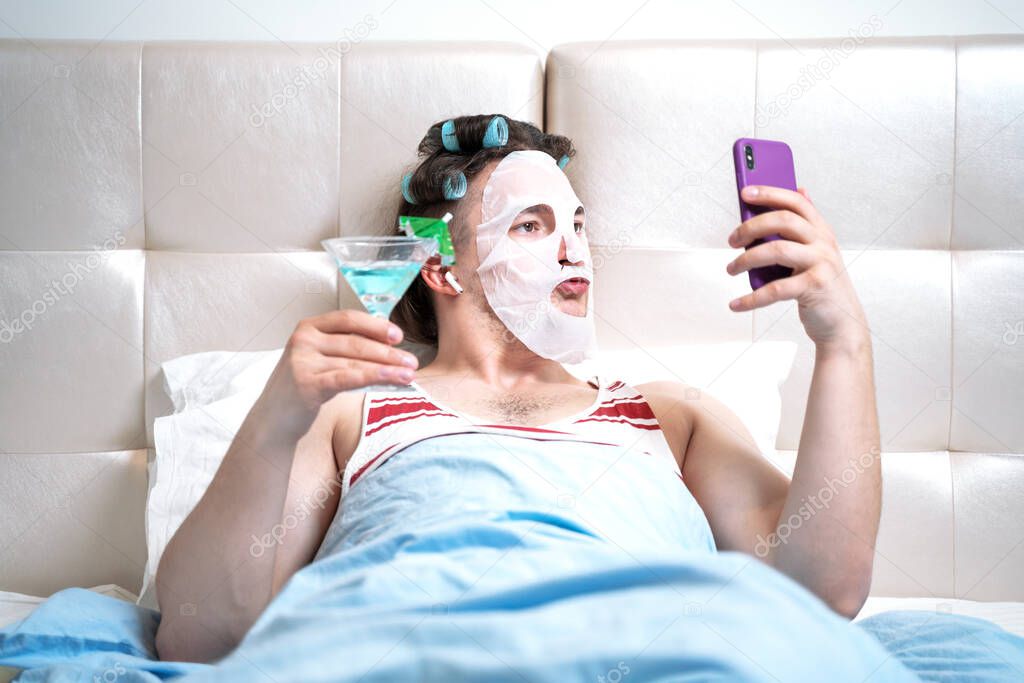 Funny man with a cosmetic mask on his face and curlers does spa treatments in a room holding a cocktail in his hand spends a stream for social networks. Parody on cosmetics beauty and fashion bloggers