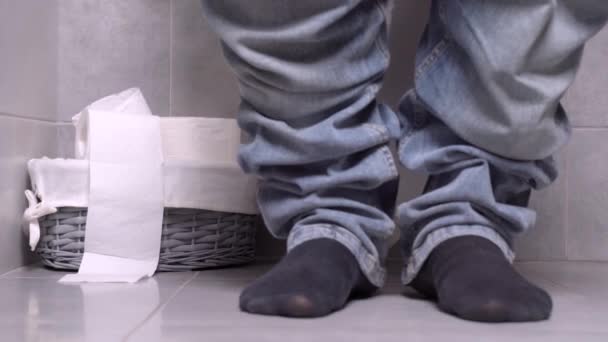 Man zit op het toilet, benen in jeans in frame. Hand reikt naar rol wc-papier in mand en scheurt stuk af. Bekende regels van persoonlijke hygiëne. Strategische voorraden kritieke goederen voor noodsituaties. — Stockvideo