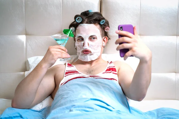 Hairy guy in striped tank top with curlers for fashionable hairstyle and with moisturizing fabric mask for wrinkles on face lies in bed. Man makes selfie on smartphone with cocktail, luxurious life.