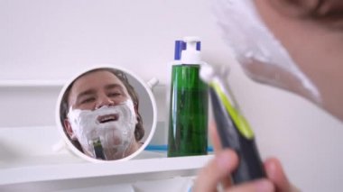 Küçük yuvarlak aynada yüzünde tıraş köpüğü olan bir adamın yansıması. Adam sakal ve bıyığı elektrikli jiletle traş ediyor. Diş fırçası ve kozmetik ürünleri banyodaki raflarda. Günlük sabah tedavileri.