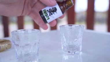 Masanın üzerinde duran iki kısa şeffaf bardak. Bir tanesinde insan eli küçük şişeden zehir damlatıyor..