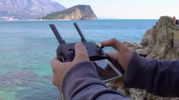 Człowiek trzyma panel sterowania dronem w rękach i przesuwa kije w lewo i prawo, zbliżenie. Telefon komórkowy jest podłączony do kontrolera, aby nadawać z kamery. Piękny krajobraz morski z górami na tle. — Wideo stockowe