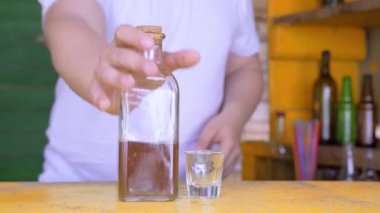 İçinde sarı sıvı bulunan şeffaf bir şişe ve masanın yanında kısa bir bardak. İnsan elleri onları alır ve geri döner..