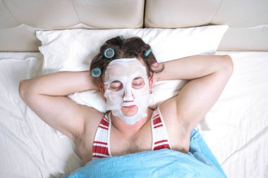 Yüzünde kozmetik maske olan genç bir adam. Gözlerinde göz yuvaları, saçlarında bigudiler, rahat bir yatakta uzanıyor, uyuyor ya da rüya görüyor..