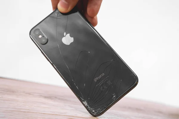 11. April 2020, Rostow am Don, Russland: Menschliche Hand hält Smartphone iPhone X von weltraumgrauer Farbe mit versehentlich zerbrochenem Rückglas mit den Fingern auf verschwommenem Hintergrund, Frontansicht, Nahaufnahme. — Stockfoto