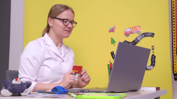 Νεαρή λογοθεραπεύτρια, γυναίκα με λευκό παλτό, που παρέχει το διαδικτυακό μάθημα χρησιμοποιώντας φορητό υπολογιστή, που δείχνει πώς να προφέρει τις λέξεις, με ισχυρή άρθρωση. Η συνεδρία για το παιδί, με τα παιδιά εικόνες γύρω — Αρχείο Βίντεο