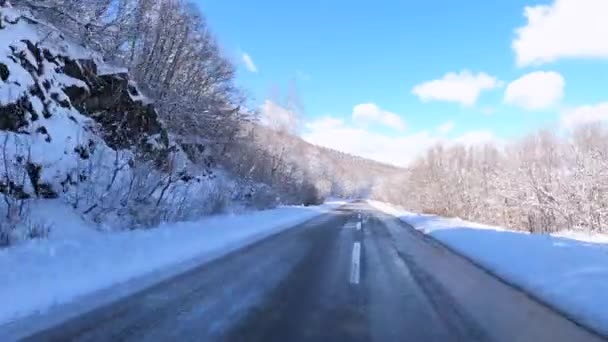 Strzelanina wykonana z przedniej szyby samochodu, poruszająca się dwupasmową drogą między zalesionymi wzgórzami pokrytymi śniegiem, gdzie nagle otwiera się wspaniały widok na góry z jasnym błękitnym niebem z chmurami. — Wideo stockowe
