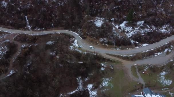 Ciężarówka jedzie krętą górską drogą węży mijając rzadkie zaśnieżone lasy, samochód zaparkowany na drodze i wioskę podgórską w chłodny wczesną wiosnę lub zimę, strzelając dronem z wysokości. — Wideo stockowe